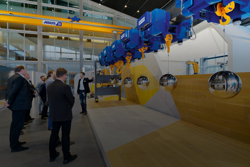 A group of visitors in the crane house is being shown how rope hoists work. The visitors are wearing formal attire. At the top in the background is a ZLK overhead crane with a load capacity of 8 tons.